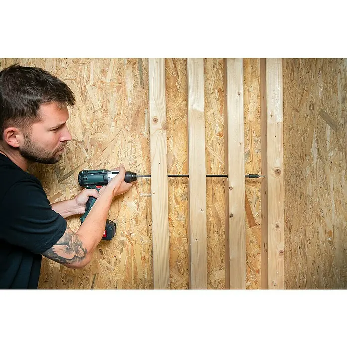 Wolfcraft Bithalter - Länge: 450 mm - Ein Mann mit Tätowierung bohrt mit einem Akku-Bohrschrauber ein langes Loch durch mehrere Holzlatten. Die Konstruktion ist eine Holzwand.