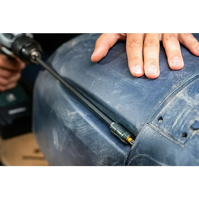 Wolfcraft Bithalter - Länge: 300 mm - Detailaufnahme eines Akkuschraubers mit langem Bit, der in ein blaues, staubiges Objekt geschraubt wird. Eine Hand liegt zur Stabilisierung auf dem Objekt.
