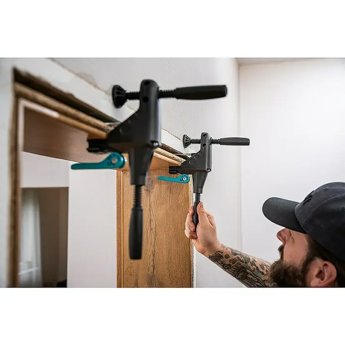 Wolfcraft Türfuttermontage-Set Pro - Passend für: Türen mit 610 - 985 mm Breite - Ein Mann befestigt eine Holztür mit zwei Korpuszwingen an der Wand. Die Zwingen sind schwarz und haben einen grünen Hebel.