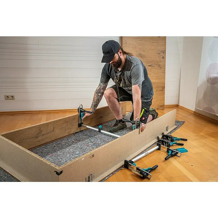 Wolfcraft Türfuttermontage-Set Pro - Passend für: Türen mit 610 - 985 mm Breite - Ein Mann fixiert mit einer Wolfcraft-Werkzeugklemme einen Holzrahmen, der auf einer schützenden Unterlage liegt.
