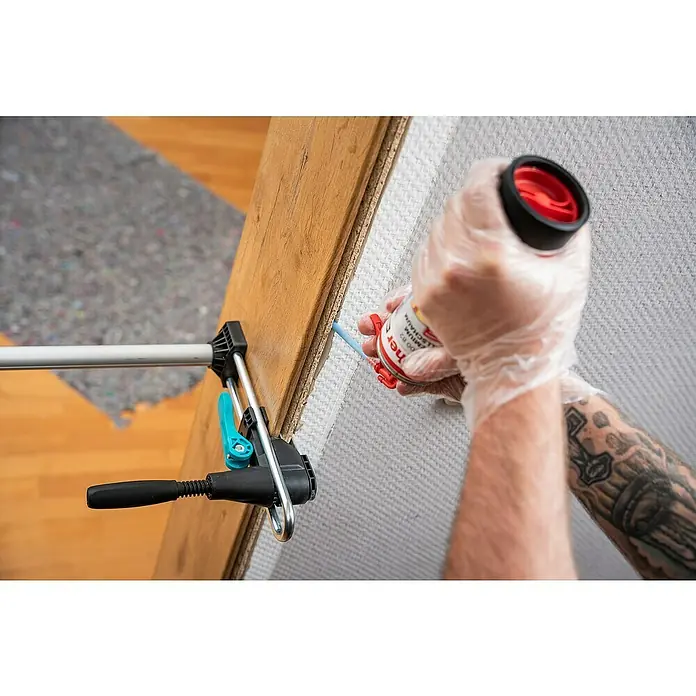 Wolfcraft Türfuttermontage-Set Pro - Passend für: Türen mit 610 - 985 mm Breite - Die Bild zeigt die Anwendung eines PU-Schaums zur Abdichtung eines Fensters mit einer Zwinge, der von einer Person mit Handschuhen aufgetragen wird.