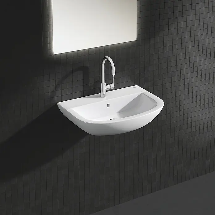 Grohe BauEdge Waschtischarmatur L-Size - Chrom, Glänzend - Ein modernes Waschbecken mit Armatur und Spiegel vor dunklem Fliesenhintergrund. Das Waschbecken ist weiß und hat eine abgerundete Form.