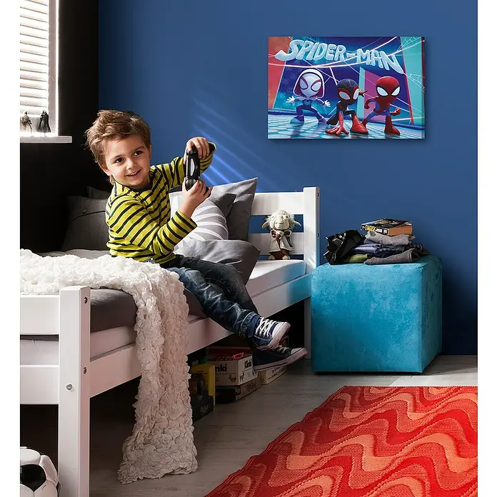 Komar Leinwandbild - Spidey & his amazing Friends - Glowy, B x H: 60 x 40 cm - Ein Junge sitzt mit einem Controller auf einem Bett, während an der Wand ein Spiderman-Poster hängt.