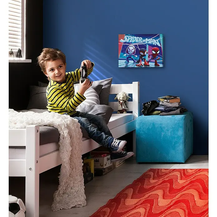 Komar Leinwandbild - Spidey & his amazing Friends - Glowy, B x H: 40 x 30 cm - Ein Junge spielt mit einem Controller in einem Zimmer mit einem Spiderman-Poster und einem blauen Hocker.