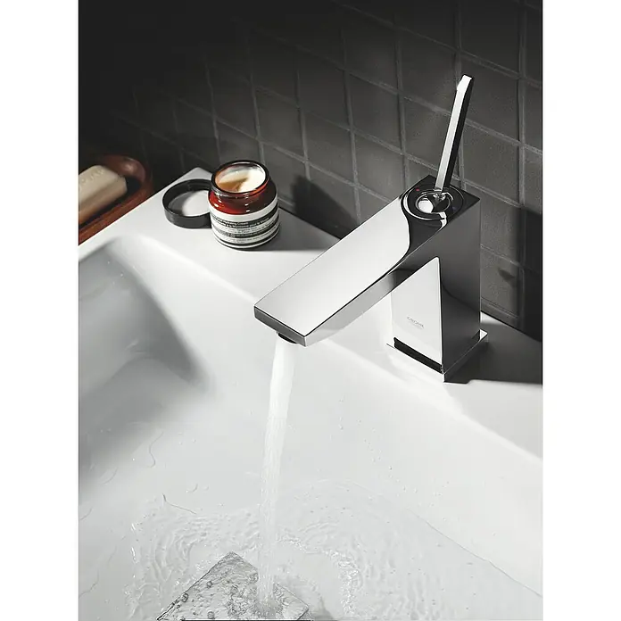 Grohe Eurocube Joy Waschtischarmatur M-Size - Chrom, Glänzend, Mit Zugstange - Nahaufnahme einer glänzenden Chrom-Armatur von Grohe mit fließendem Wasser in einem Waschbecken, daneben ein offener Tiegel Hautcreme.