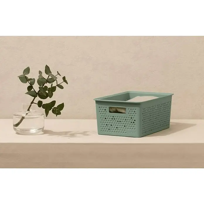 Tatay Bohol Cesta para baño - L x An x Al: 17,5 x 27 x 11,5 cm, Verde sage - Primer plano de una cesta verde menta que contiene ropa, sentada sobre una superficie blanca junto a un recipiente de vidrio con una planta.