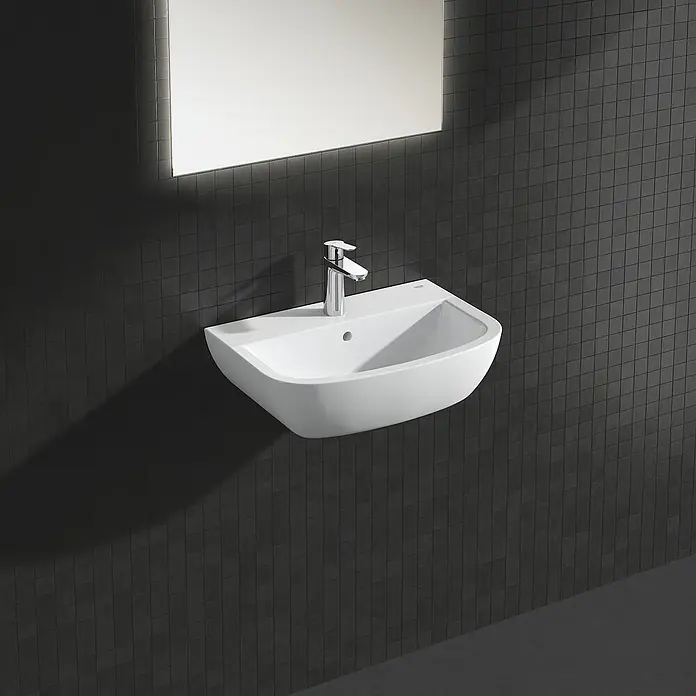 Grohe BauEdge Waschtischarmatur M-Size - Chrom, Glänzend - Ein modernes Waschbecken aus weißer Keramik mit einem chromfarbenen Wasserhahn, das an einer dunklen, gefliesten Wand montiert ist und über einen beleuchteten Spiegel verfügt.