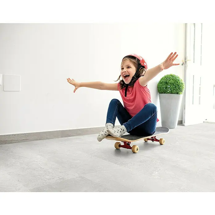 Decolife Designboden Naturestyle Pure - Slate Light Grey, Fliese, 905 x 295 x 9 mm - Ein fröhliches Mädchen mit Helm fährt auf einem Skateboard in einem hellen Raum, die Arme ausgebreitet, und strahlt pure Freude aus.
