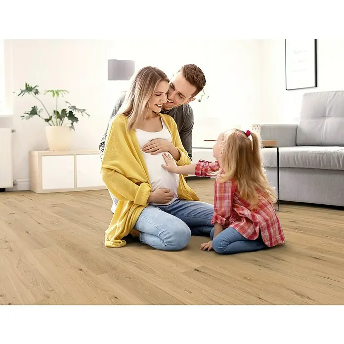 Decolife Designbodenmuster Naturestyle Pure Oak Sun - 28 x 16 x 9 mm, Landhausdiele, Oak Sun - Ein helles Holzlaminatboden ist in einem hellen Raum verlegt, in dem eine schwangere Frau mit ihrer Familie sitzt.