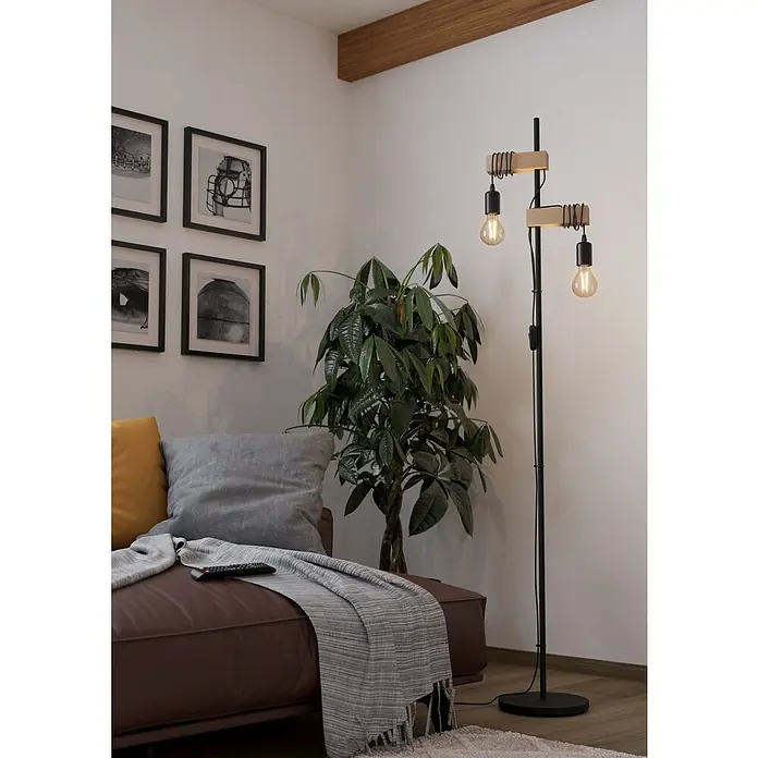 Eglo Townshend Stehleuchte - 20 W, Höhe: 166,5 cm, Braun/Schwarz, E27 - Eine moderne Stehlampe mit zwei Glühbirnen und Holzelementen steht neben einer Zimmerpflanze und einem Sofa im Wohnzimmer.