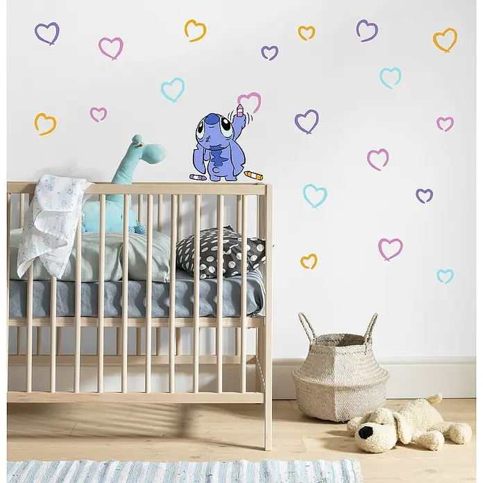 Komar Wandtattoo Stitch Artist - Bunt, 22 -tlg. - Ein Kinderzimmer mit einem Babybett, einer Stofftierpuppe und einem Stoffkorb, dekoriert mit bunten Herzaufklebern an der Wand.