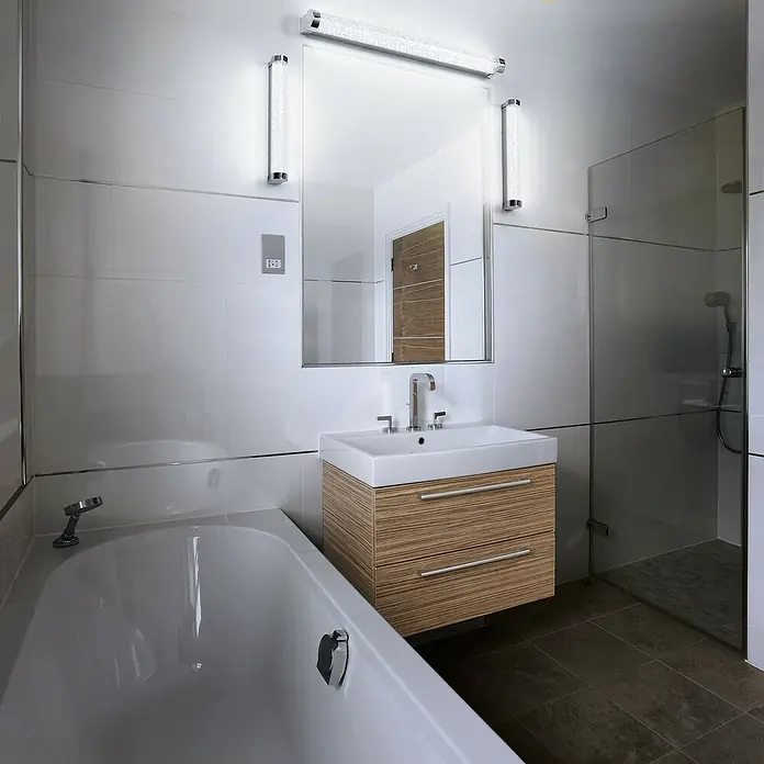 Ledvance Orbis LED-Spiegelleuchte Tube - 12 W, L x B x H: 60 x 7 x 5,4 cm, Chrome, Bubbleglas, Warmweiß - Modernes Badezimmer mit weißer Badewanne und Waschtisch mit Holzfront, beleuchtetem Spiegel und Dusche mit Glastrennwand.