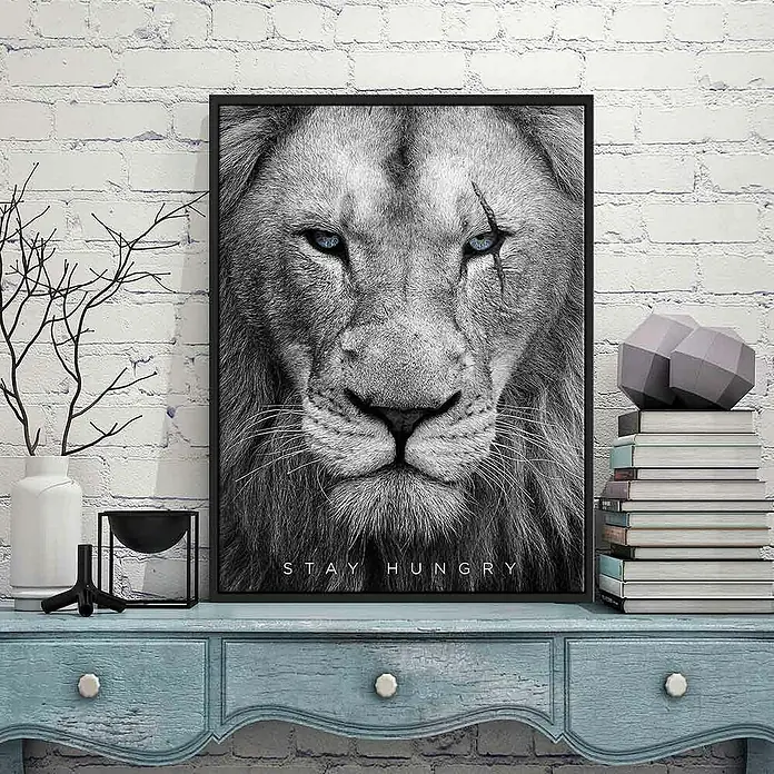 Dotcomcanvas Leinwandbild - Hungry Face, B x H: 60 x 80 cm - Ein gerahmtes Löwenporträt in Schwarzweiß mit blauen Augen und dem Schriftzug „Stay Hungry“, das eine auffällige Wanddekoration darstellt.