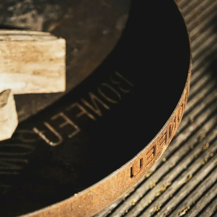 Bonfeu Brasero Bonbowl Plus - Diámetro: 60 cm, Acero Corten - Primer plano de un disco metálico redondo y oxidado con letras grabadas en una superficie texturizada.