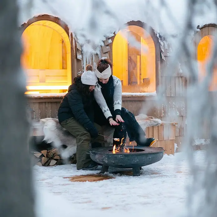 Bonfeu Brasero Bonfeu Plus - Diámetro: 80 cm, Acero Corten - Dos personas asan malvaviscos sobre una fogata en la nieve, frente a una cabaña de madera iluminada. Escena invernal con romanticismo.