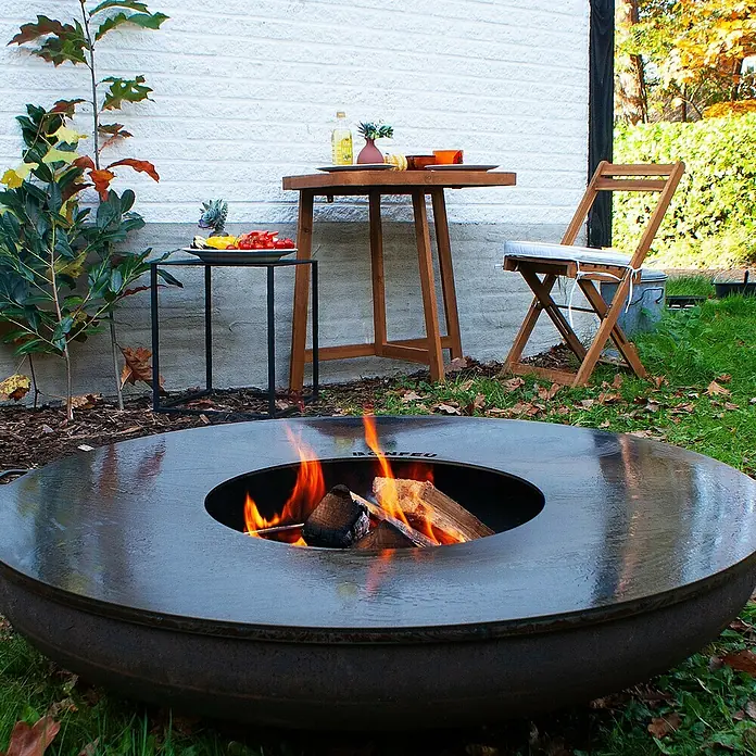 Bonfeu Brasero Bonfeu Plus - Diámetro: 80 cm, Acero Corten - Ambiente exterior con un brasero Bonfeu, que proporciona calor y ambiente a un espacio de jardín, complementado con muebles rústicos.