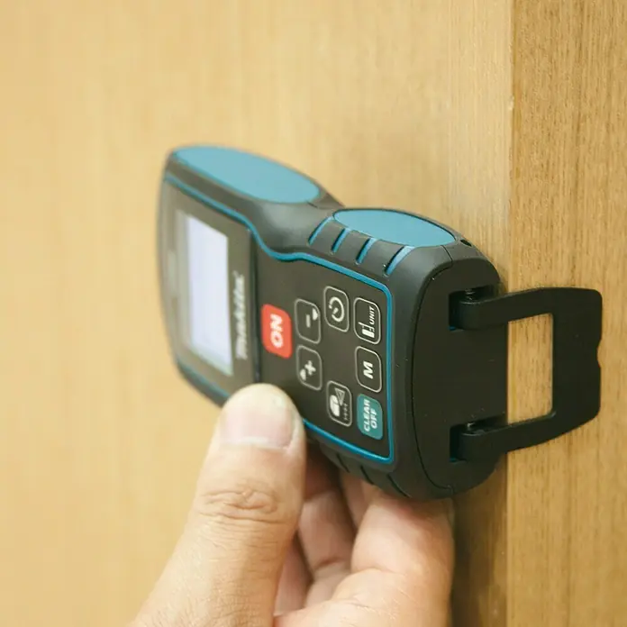 Makita Laserentfernungsmesser LD080P - Messbereich: 0,05 - 100 m - Nahaufnahme eines Makita-Laser-Entfernungsmessers, das an einer Holzkante befestigt ist und von einer Hand gehalten wird, um präzise Messungen zu gewährleisten.