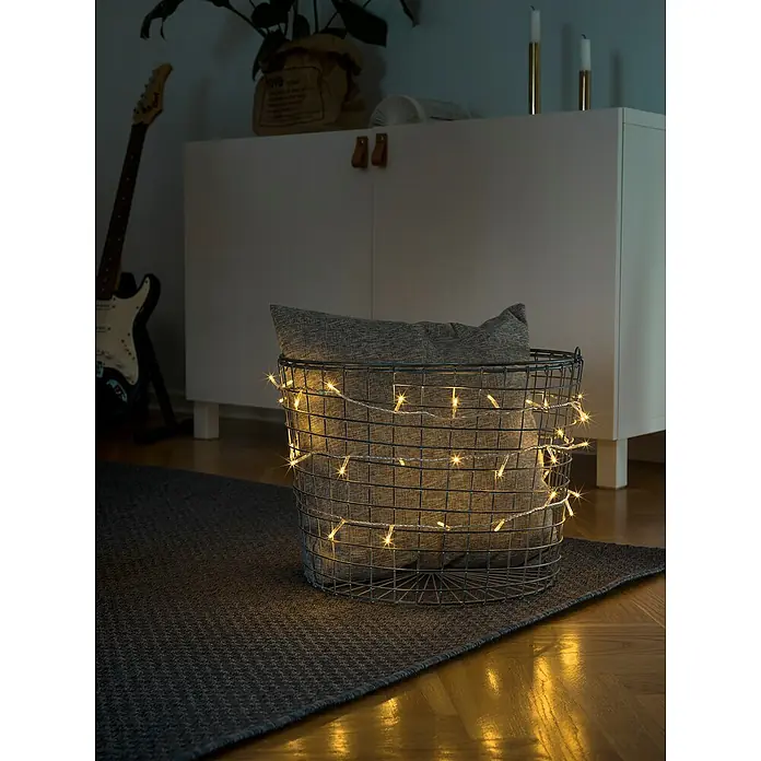 Konstsmide LED-Lichterkette - Innen, Lichtfarbe: Warmweiß, Anzahl Leuchtmittel: 35 Stk., Netzbetrieben, Transparent - Nahaufnahme eines Drahtkorbs mit Kissen, der mit warmweißen LED-Lichtern umwickelt ist, die ein gemütliches Ambiente schaffen.