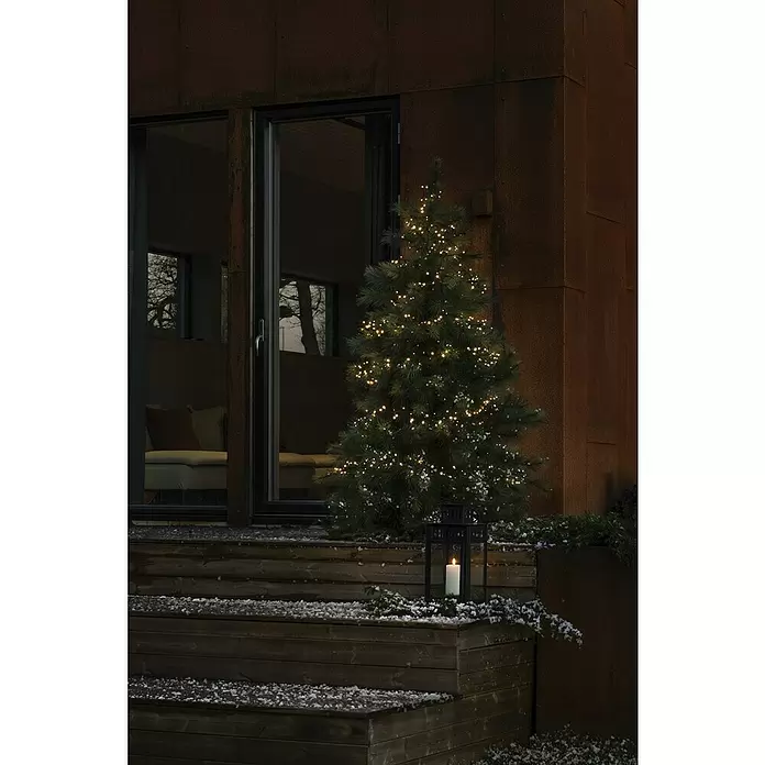 Konstsmide LED-Lichterkette - Außen, Lichtfarbe: Warmweiß, Anzahl Leuchtmittel: 600 Stk., Netzbetrieben - Ein Weihnachtsbaum mit warmweißen Lichtern auf verschneiten Holzstufen, neben einer schwarzen Laterne mit Kerze.