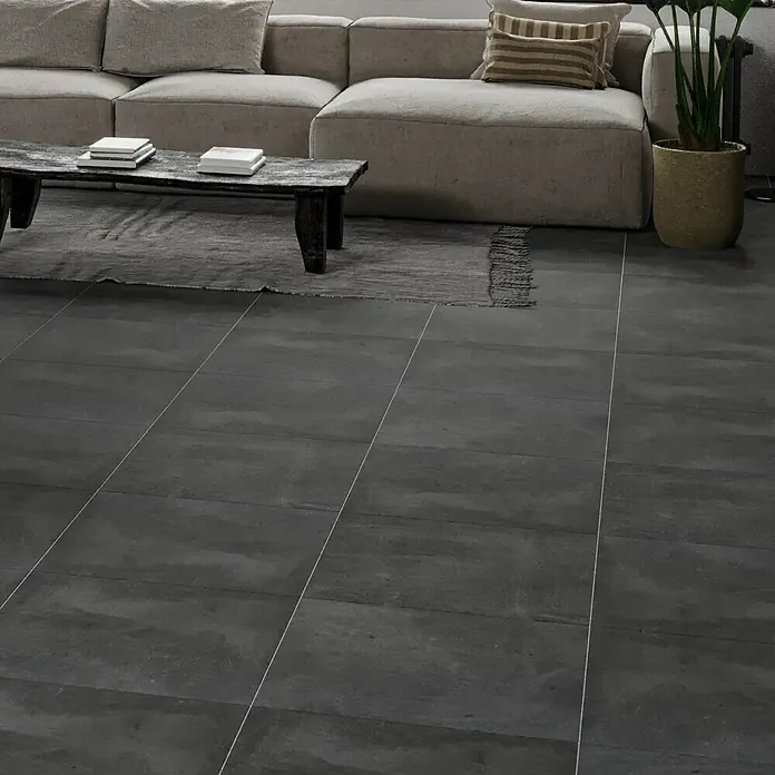 Feinsteinzeugfliese Manhattan Dark - 60 x 60 cm, Anthrazit, Matt - Ein modernes Wohnzimmer mit grauen Fliesen, einem bequemen Sofa und einem dunklen Holztisch. Die Fliesen verleihen dem Raum eine elegante Note.