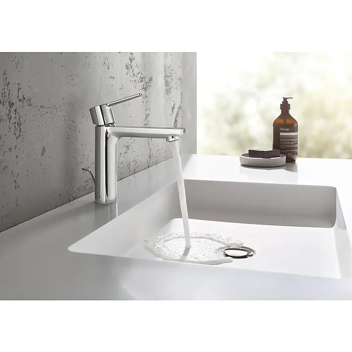 Grohe Lineare Waschtischarmatur S-Size - Mit Zugstange, Chrom - Nahaufnahme eines modernen Chrom-Wasserhahns in einem Waschbecken, aus dem Wasser fließt, mit einer Seife und Lotion im Hintergrund.