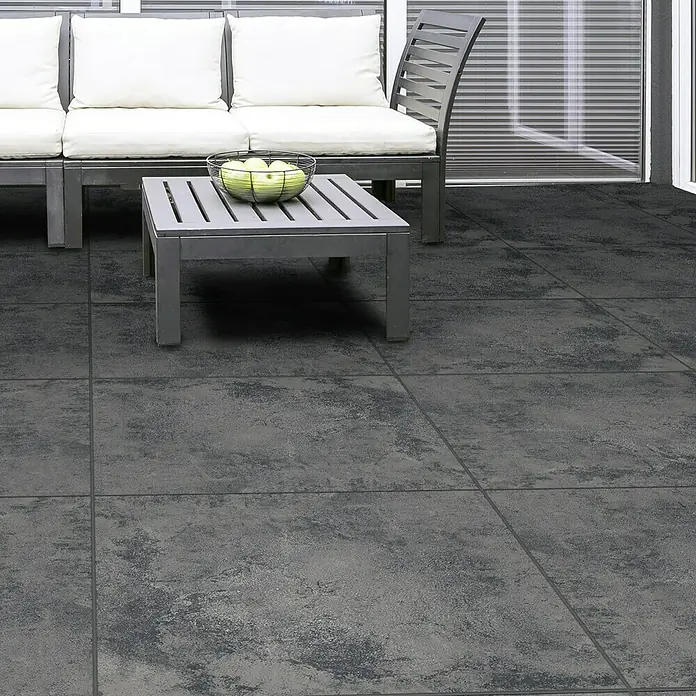 Queens Terrassenfliese - 59,3 x 59,3 x 2 cm, Graphit, Matt - Nahaufnahme eines grauen Terrassenbereichs mit Fliesenboden, Couch, Tisch und einer Schale mit grünen Äpfeln.