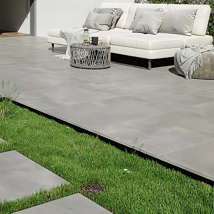 Queens Terrassenfliese - 59,3 x 59,3 x 2 cm, Grau, Matt - Ein stilvolles Outdoor-Ambiente mit grauem Terrassenboden, ergänzt durch ein weißes Sofa mit Kissen und Beistelltisch.