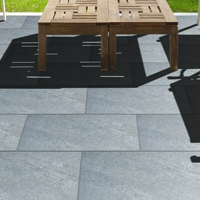 Terrassenfliese Nevada - 60 x 60 x 2 cm, Grau, Matt - Außenaufnahme von grauen Pflastersteinen, die eine Terrasse bilden, mit einem Teil eines Holztisches, der darauf Schatten wirft.