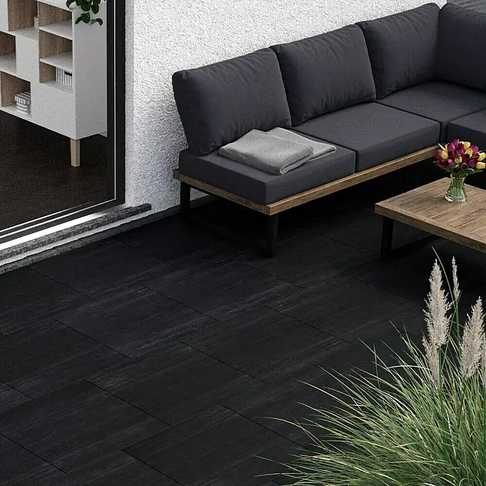 Terrassenfliese Garden Black Struktur - 59,7 x 59,7 x 2 cm, Schwarz, Matt - Eine stilvolle Außenterrasse mit schwarzen Fliesen, die einen modernen Loungebereich mit grauer Polsterung und Holztisch ergänzen. Die dunklen Fliesen erzeugen eine elegante Atmosphäre.