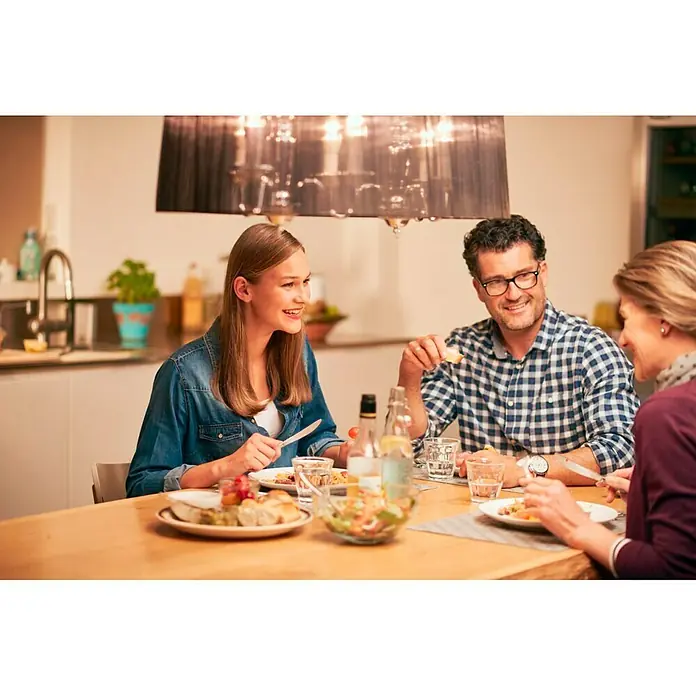 Philips Bombilla LED Vela - E14, Intensidad regulable, Blanco cálido, 470 lm, 6 W - Tres personas están sentadas en la mesa del comedor comiendo juntas. Una lámpara cuelga sobre la mesa.