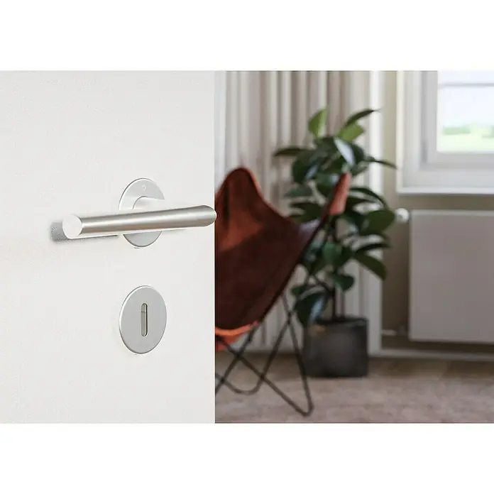 Hoppe Zimmertürgarnitur Stockholm - Stärke Tür: 37 mm - 42 mm, Aluminium, Buntbart - Nahaufnahme eines Türgriffs aus Metall an einer weißen Tür. Dahinter ein Zimmer mit Sessel und Topfpflanze.
