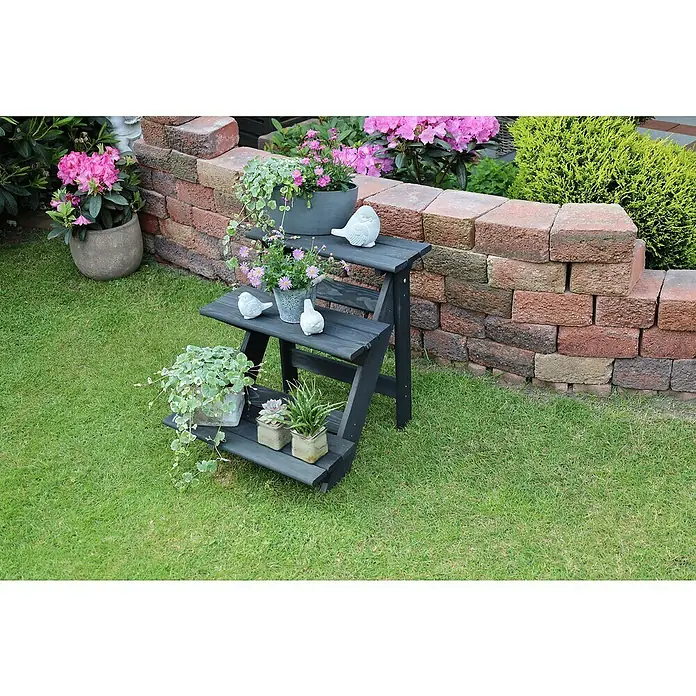 Promadino Blumentreppe - 60 x 50 x 56 cm, Kiefer, Anthrazit - Eine dreistufige, schwarze Holzleiter dient als Pflanzenständer im Garten, dekoriert mit Blumen und Vogelstatuen.
