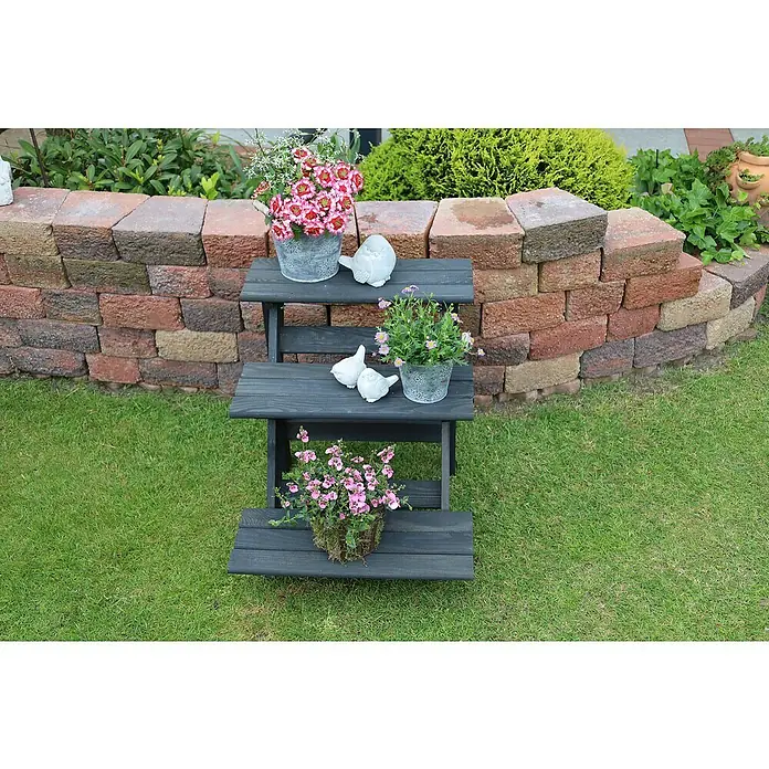 Promadino Blumentreppe - 60 x 50 x 56 cm, Kiefer, Anthrazit - Dreistufiges Pflanzenregal aus dunklem Holz, dekoriert mit Blumen und Vogelstatuen, vor einer Gartenmauer.