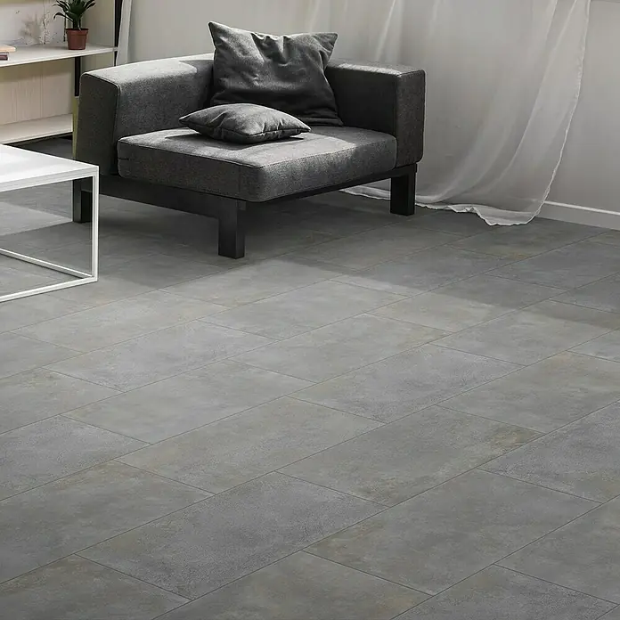 Feinsteinzeugfliese Metallbeton - 30 x 60,4 cm, Hellgrau, Matt - Ein grauer Sessel mit Kissen auf einem grauen Fliesenboden neben einem weißen Tisch und einem Regal. Moderne Inneneinrichtung mit schlichtem Design.