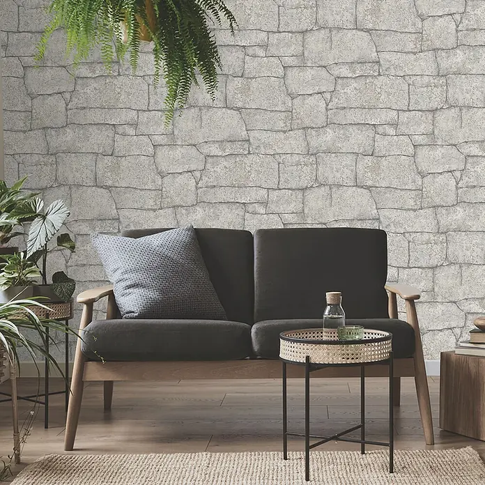 Papel pintado Muro de piedra - Blanco, 10 x 0,53 m - Primer plano de una habitación con un sofá, una mesa con una botella de agua y una planta al fondo frente a un papel tapiz de pared de piedra.