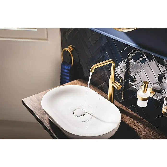 Grohe Essence New Waschtischarmatur XL-Size - Cool Sunrise, Matt - Eine Nahaufnahme eines Waschbeckens mit goldener Armatur, auf dem Wasser läuft, Teil einer eleganten Badezimmerausstattung.