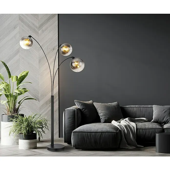 Globo Newcastle Stehleuchte - 60 W, Höhe: 200 cm, Schwarz matt, Rauch - Elegante Stehlampe mit drei Armen und Glaskugeln, die ein warmes Licht verbreiten, neben einem grauen Sofa und Pflanzen.