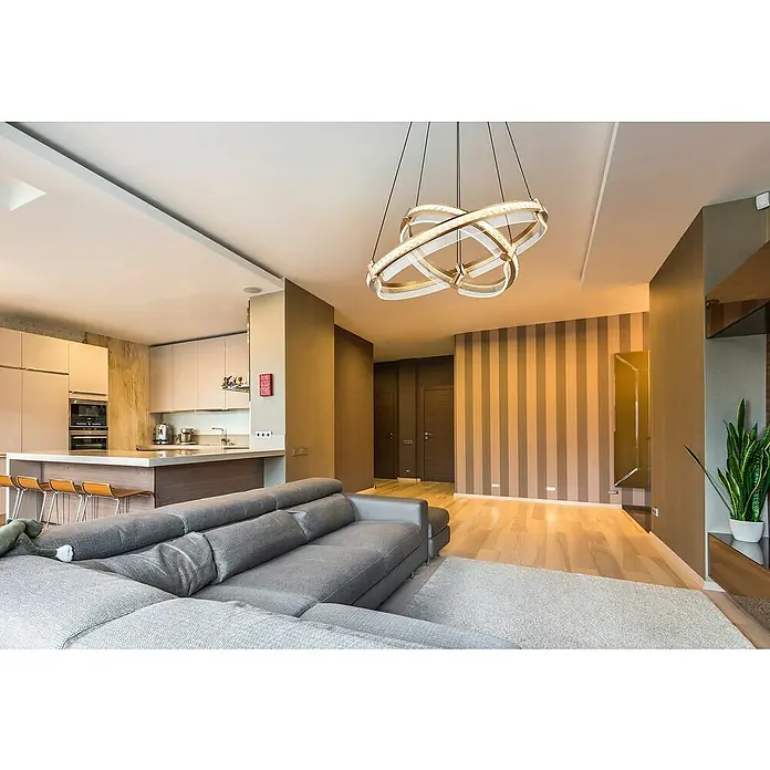 Globo Blondie LED-Pendelleuchte - 78 W, 60 x 120 cm, Gold, Gold, Mehrfarbig - Moderne Wohnzimmeransicht mit auffälliger, kreisförmiger Hängeleuchte über einer grauen Couch und einer offenen Küche im Hintergrund.