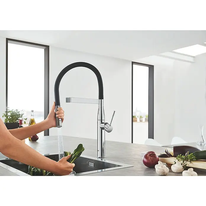 Grohe Essence Spültischarmatur - Chrom, Glänzend - Eine verchromte GROHE Küchenarmatur mit hohem Auslauf wird benutzt, um Gemüse über einer Spüle in einer hellen Küche zu waschen.