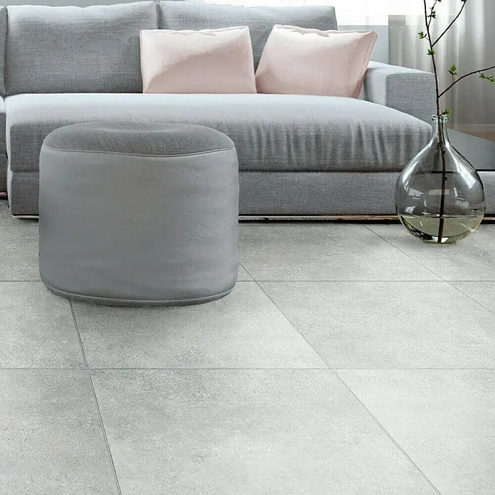 Feinsteinzeugfliese Beton - 60 x 60 cm, Grau, Matt - Grauer Hocker aus Stoff vor einem Sofa mit Kissen und einer Glasvase mit Zweigen im Wohnzimmer auf Fliesen.