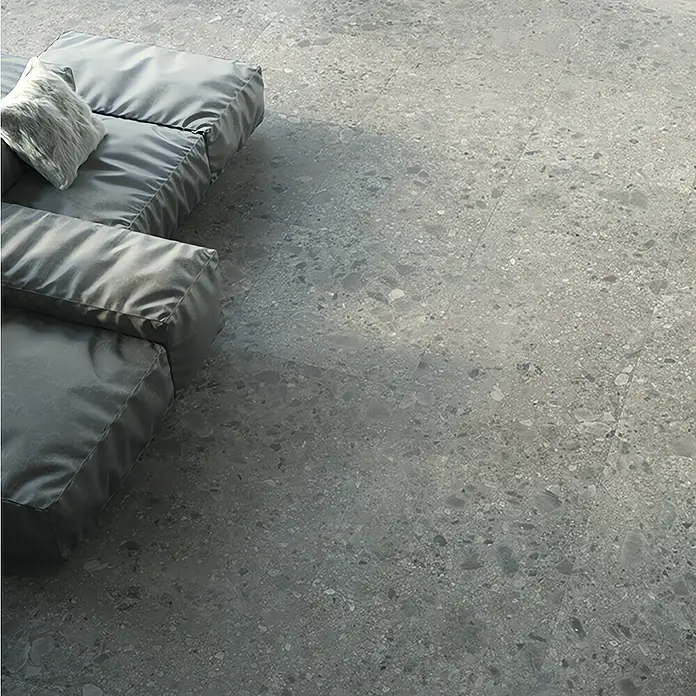 Feinsteinzeugfliese Hannover Steel - 60 x 60 cm, Grau, Matt - Grauer Terrazzoboden mit Sitzkissen. Das Bild zeigt die nahtlose Eleganz eines modernen Terrazzobodens mit einem stilvollen grauen Sitzkissen.