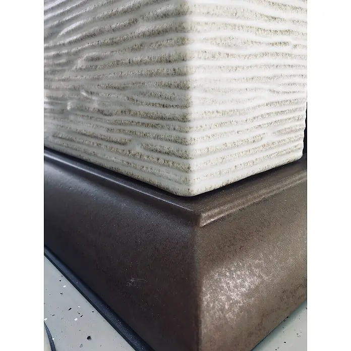 Domofire Kaminofen Cortina - 8 kW, Raumheizvermögen: 160 m³, Material Verkleidung: Keramik, Cremebeige-Tabakbraun - Nahaufnahme einer quadratischen Tischlampe mit horizontal gerippter Textur auf einem abgestuften Sockel mit Metallic-Finish.