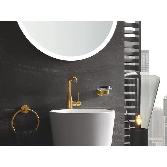 Grohe Essence New Waschtischarmatur L-Size - Mit Zugstange, Cool Sunrise, Matt - Elegantes Badezimmer-Interieur mit einem runden Spiegel, goldener Armatur und passenden Accessoires. Waschbecken und Lampe ergänzen das luxuriöse Design.