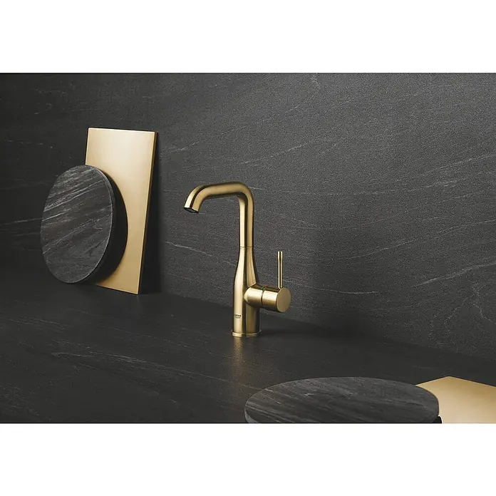 Grohe Essence New Waschtischarmatur L-Size - Mit Zugstange, Cool Sunrise, Matt - Nahaufnahme einer goldenen Grohe Armatur auf einem dunklen Waschtisch mit dekorativen Elementen. Elegantes Design für moderne Badezimmer.