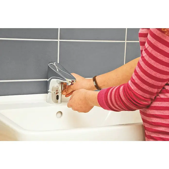 Grohe Europlus Waschtischarmatur mit Temperaturregulierung, batteriebetrieben - Infrarot-Elektronik, Chrom - Eine Person wäscht sich die Hände an einem modernen, verchromten Wasserhahn mit Sensorfunktion in einem Badezimmer mit grauen Fliesen.