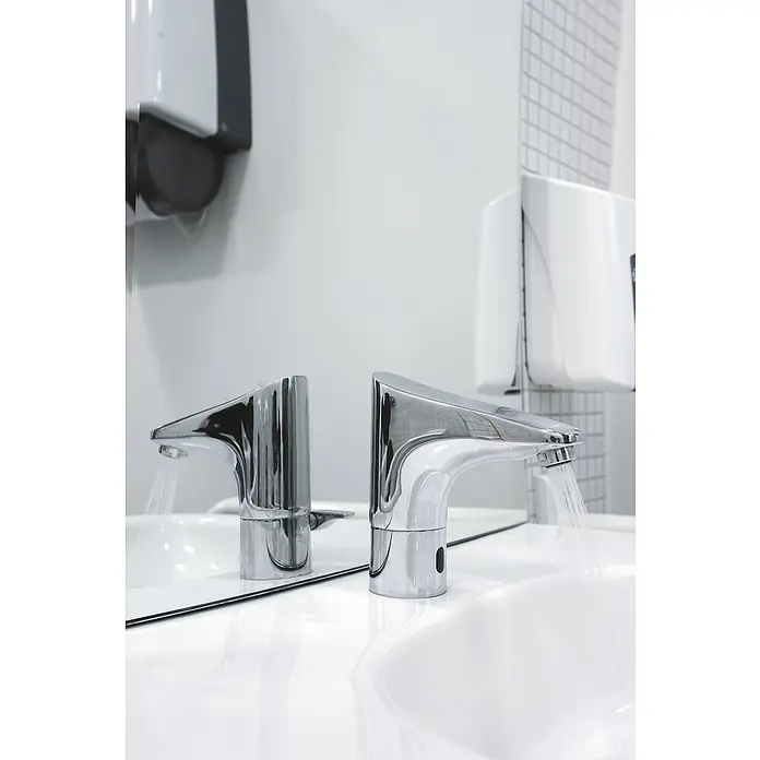 Grohe Europlus Waschtischarmatur mit Temperaturregulierung, batteriebetrieben - Infrarot-Elektronik, Chrom - Zwei moderne, berührungslose Chromarmaturen auf einem weißen Waschbecken mit fließendem Wasser, daneben Seifen- und Händetrockner.