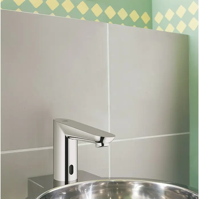 Grohe Euroeco CT Waschtischarmatur netzbetrieben - Infrarot-Elektronik, Chrom - Nahaufnahme einer modernen, berührungslosen Armatur mit Chromoberfläche vor einem Hintergrund aus grauen Fliesen und einer Wand mit Rautenmuster.