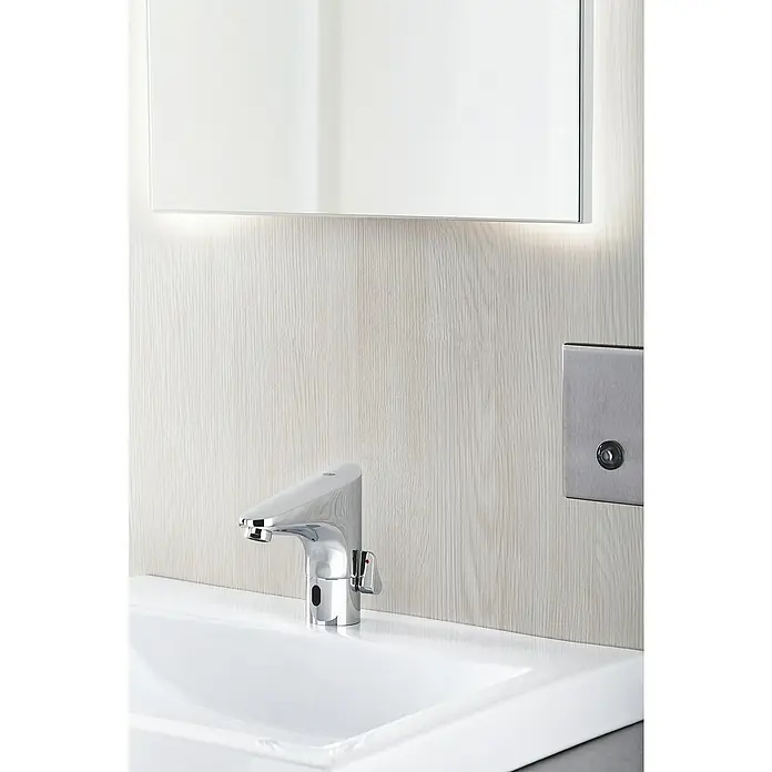 Grohe Europlus Waschtischarmatur mit Temperaturregulierung, batteriebetrieben - Infrarot-Elektronik, Chrom - Nahaufnahme eines modernen, verchromten Wasserhahns am Waschbecken mit beleuchtetem Spiegel und Paneel an der Wand.