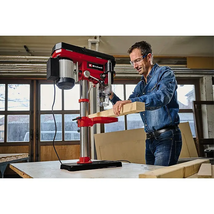 Einhell Tischbohrmaschine TC-BD 630/1 - 630 W, Leerlaufdrehzahl: 250 U/min - 2.400 U/min, Spannweite Bohrfutter: 1,5 mm - 16 mm - Ein Mann, der eine Einhell Tischbohrmaschine benutzt, um ein Stück Holz zu bohren. Die Maschine ist rot und schwarz.
