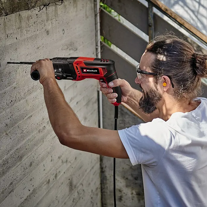 Einhell Classic Kombihammer TC-RH 800 4F - 800 W, 2,6 J, SDS-Plus-Aufnahme - Mann bohrt mit einer roten Einhell Schlagbohrmaschine ein Loch in eine Betonwand, mit Schutzbrille und Ohrstöpsel.